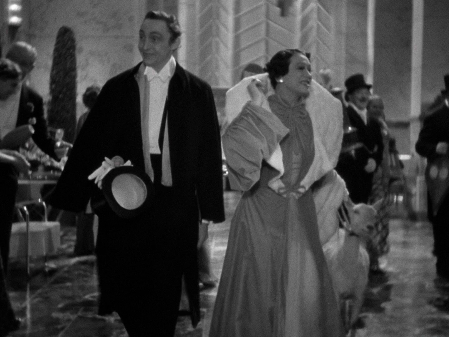 An ankles up shot of a foolish lookin man in white tie and tails, carrying a top hat and gloves, following Alice Brady across a marble-floored ballroom. Brady wears an absolute boat of a dress and is leading a baby goat on a leash. Other revelers are visible behind them.