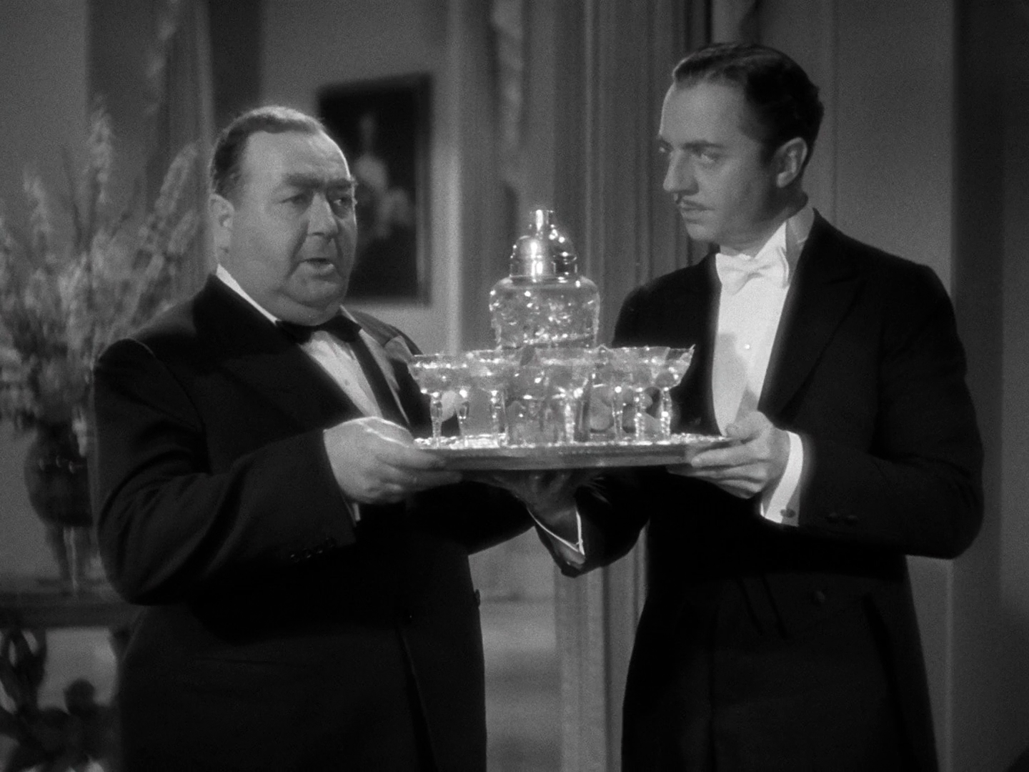 A medium shot of Eugene Pallette, in black tie, taking a silver tray of at least eight martinis, in cut crystal glasses, and a cut crystal shaker with more, from William Powell, in white tie.