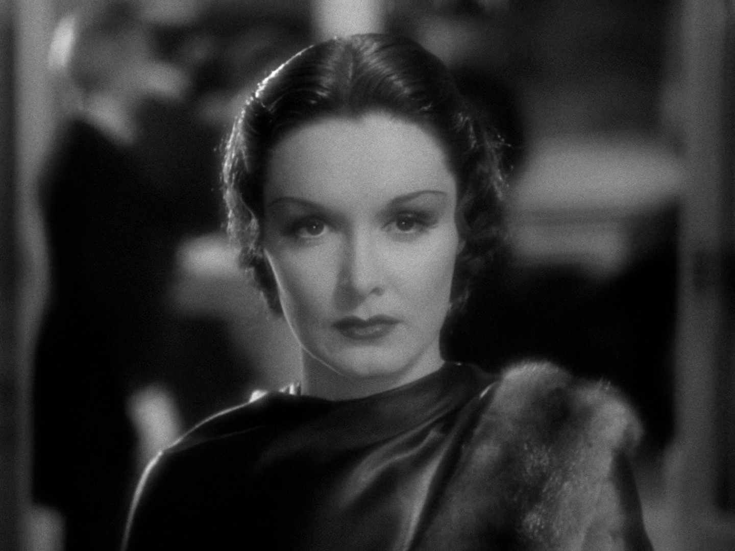 A medium close up of Gail Patrick looking right into the camera. Her dark hair is parted in the middle and is as shiny as her dress, which is also dark and seems to be silk or satin. She is not happy in this shot but the overall effect is smoldering.