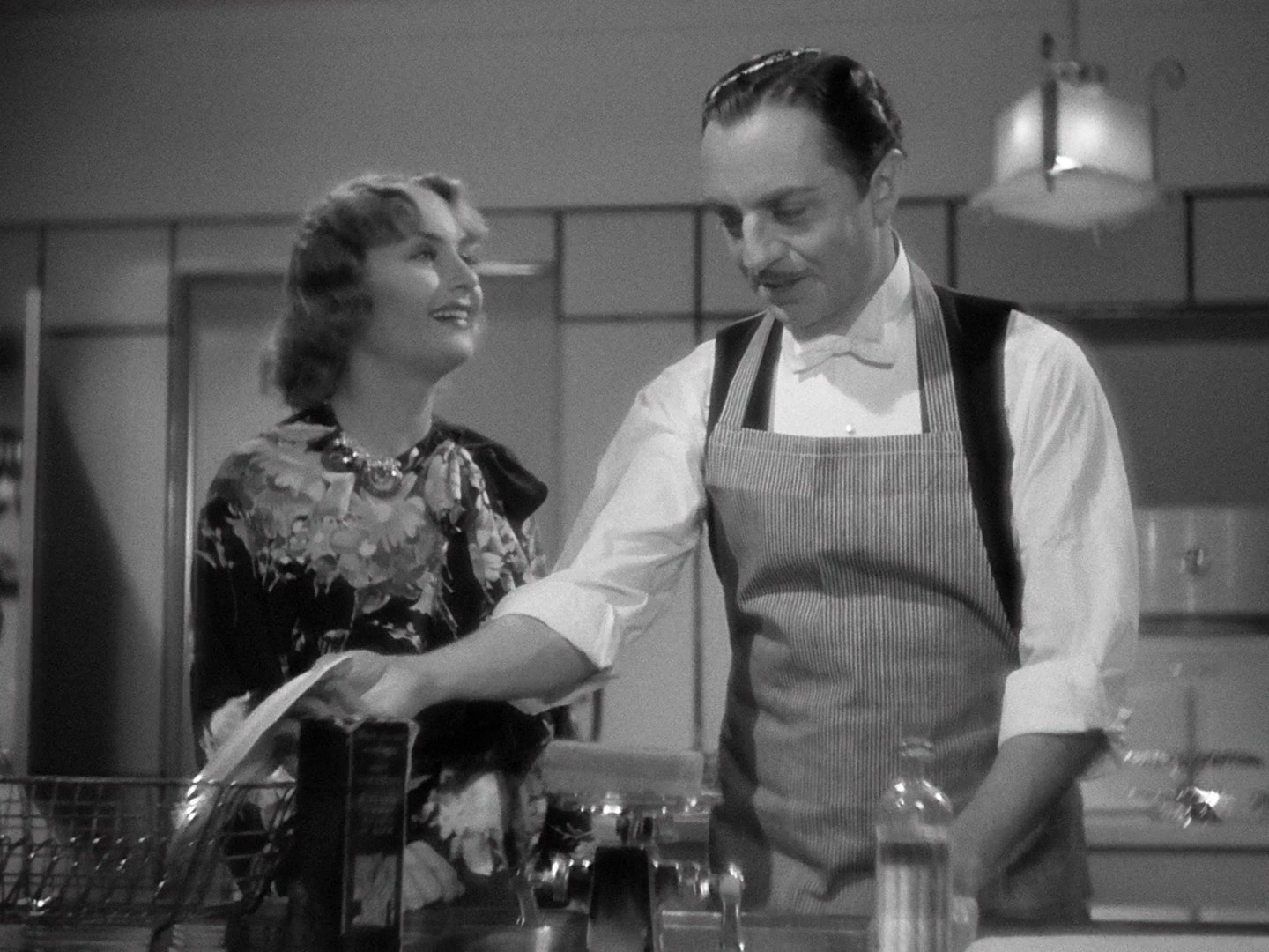 A medium shot of Carole Lombard and William Powell in a kitchen; in the foreground bottom of the frame are a dish drying rack, a kitchen sink, and a bottle of detergent. Lombard, on the left, is in a flower print dress; her shoulders are angled toward Powell and she is smiling with adoration. Powell, to the right, wears a white tie shirt and vest with a vertically striped apron, and is looking amused as he sets a dish in the rack.