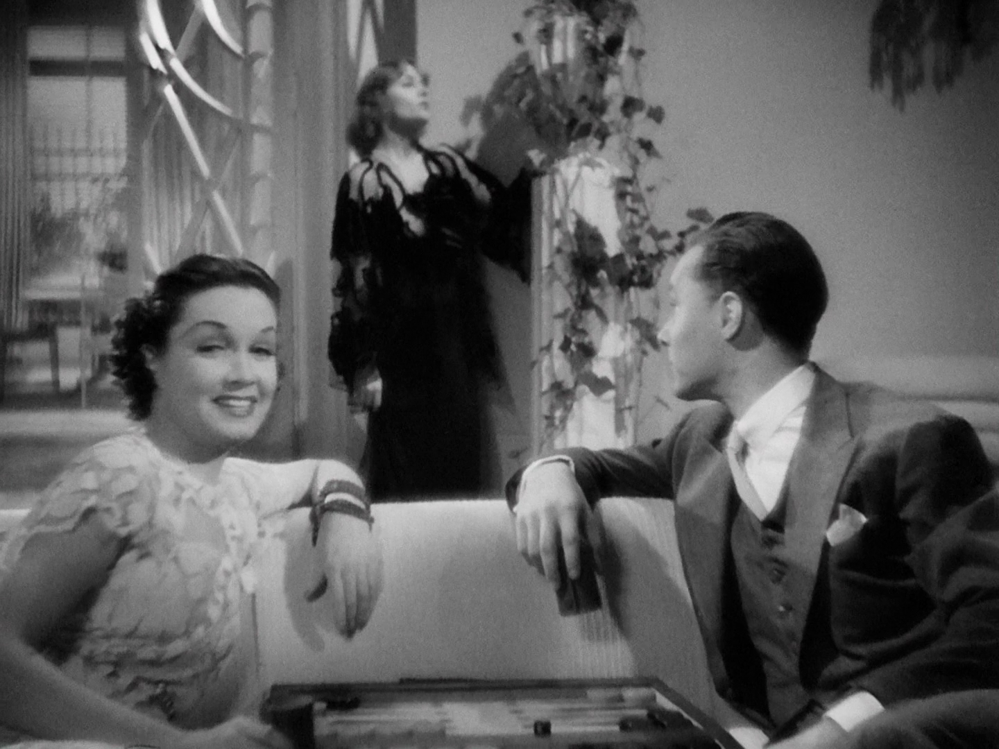 A deep focus shot of a living room. In the foreground, a couch crosses the frame; Gail Patrick, in a white lace dress, sits on the left and Robert Light, in a three-piece suit, sits on the right, with a backgammon board in the extreme foreground. Patrick's left hand rests on the back of the couch, as does Light's left hand. Patrick looks at the camera, smiling. Between and behind them, Carole Lombard, in a black lace dress, visible from the knees up, is standing in a woeful pose, with her hand on a column near the back wall. 