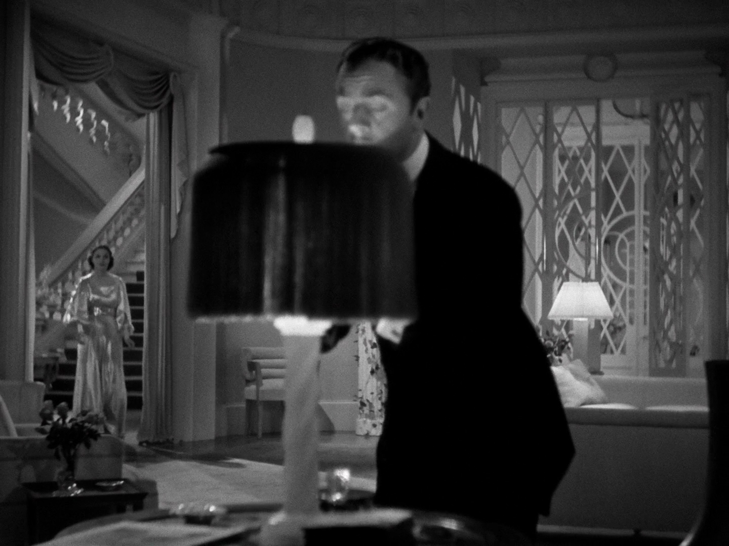 A medium shot of Wiliam Powell, in his butler white-tie, turning on a table lamp with a dark shade in the extreme foreground. Gail Patrick is in the background to the left, visible through an open doorway.